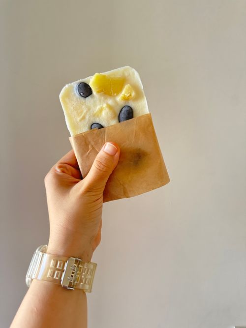 Free A hand holds a blueberry pineapple popsicle in brown paper, against a neutral backdrop. Stock Photo