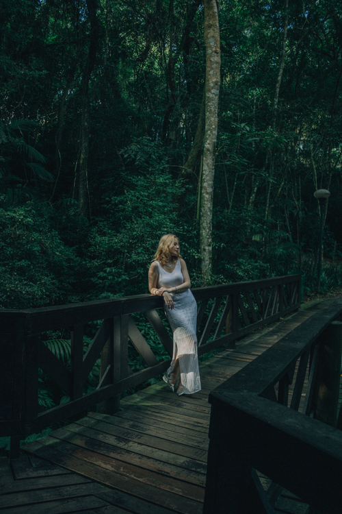 Free Elegant woman in a long dress stands on a wooden bridge in a lush forest setting. Stock Photo