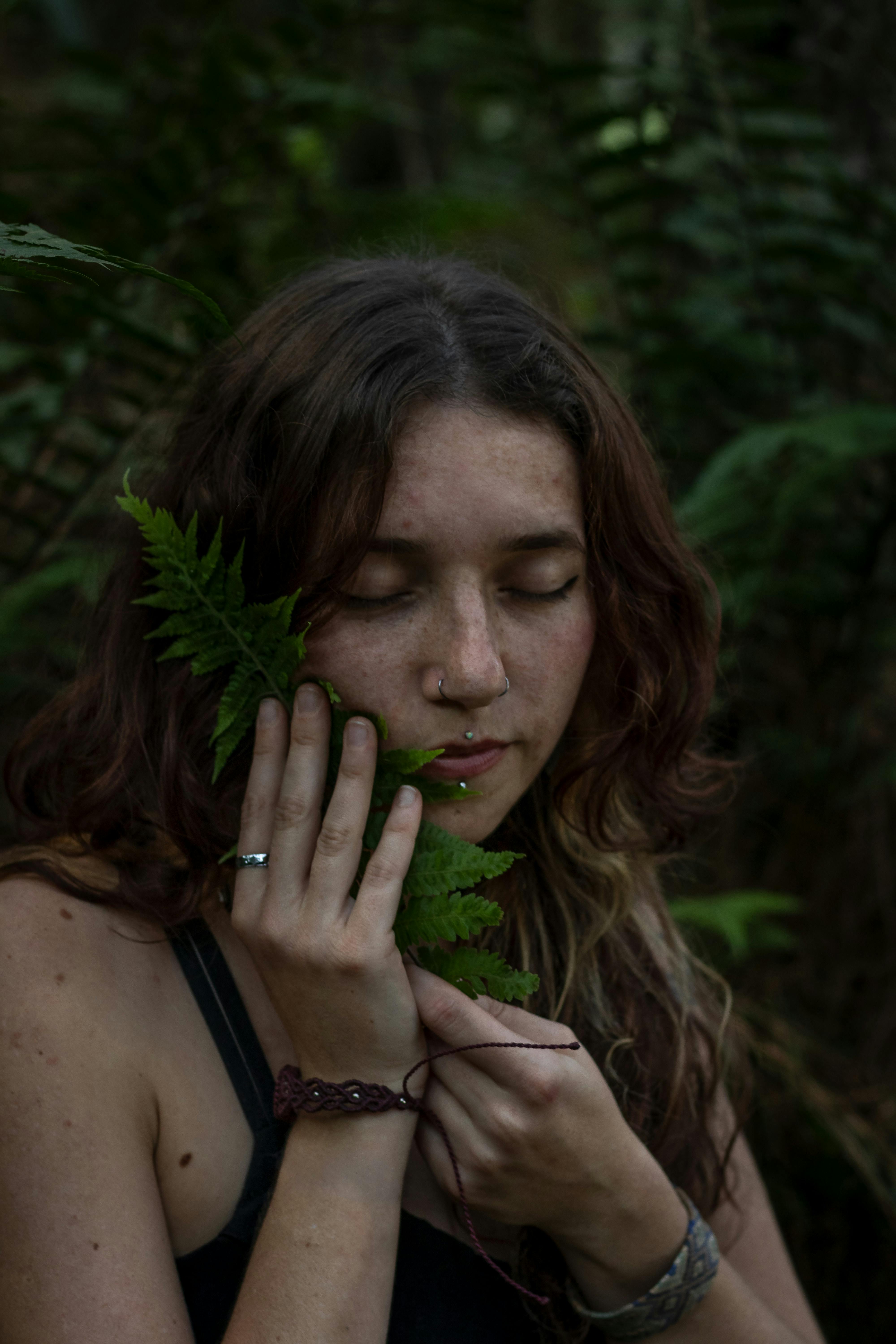 A woman closes her eyes while holding a fern leaf, surrounded by lush forest greenery.