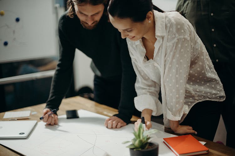 Man And Woman Leaning On Table Staring At White Board On Top Of Table Having A Meeting