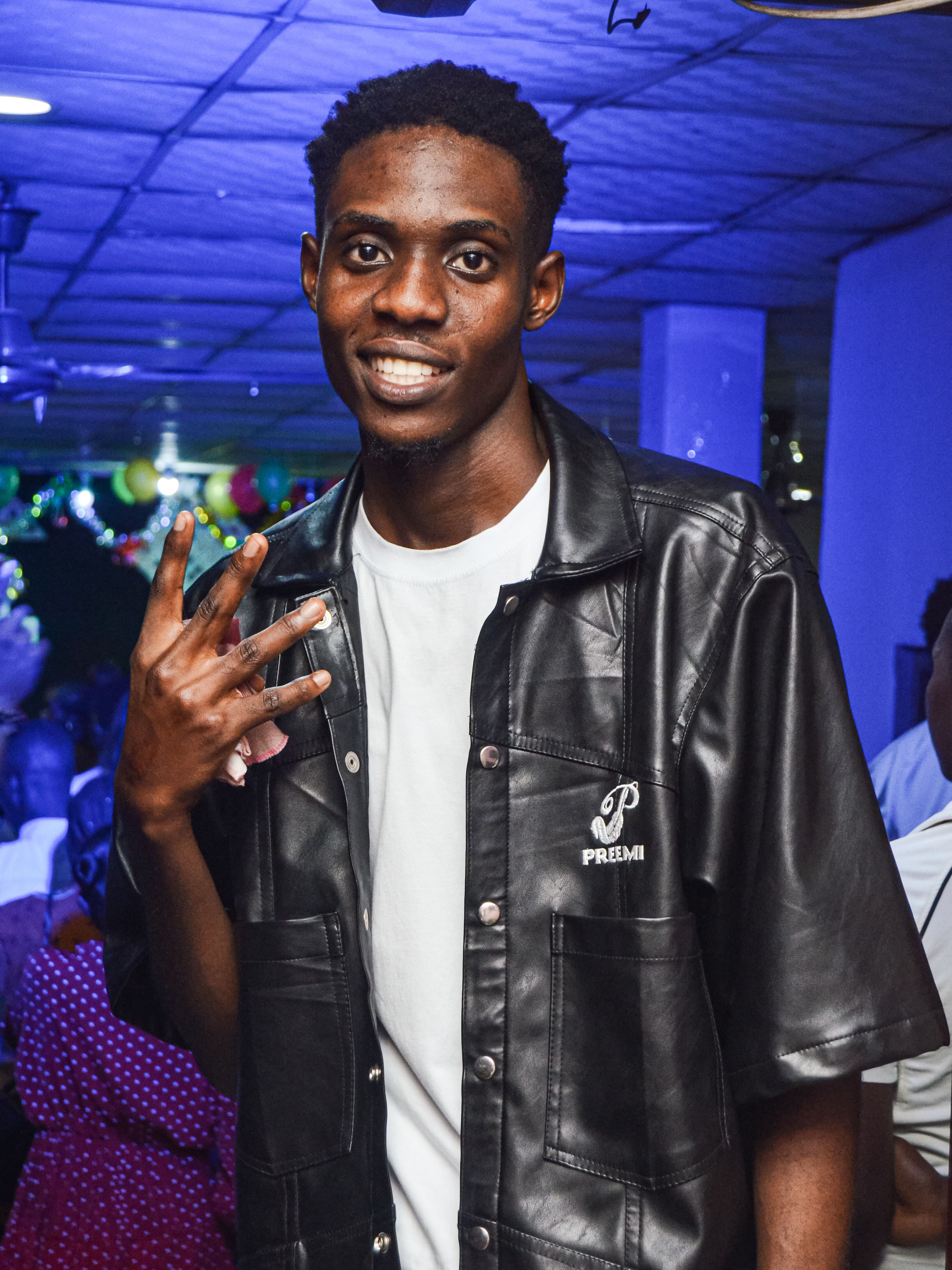 Stylish young man posing at a lively indoor Nigerian party, showcasing current fashion trends.