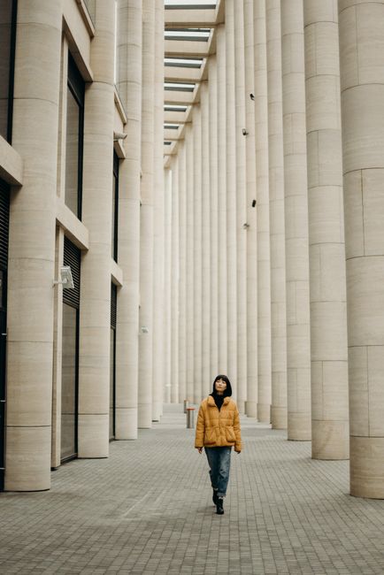 A woman in a yellow jacket strolls through a modern architectural setting with tall columns.