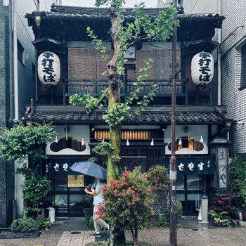 Rain-soaked day in front of a traditional Japanese restaurant with a passerby under an umbrella.（イメージ）
