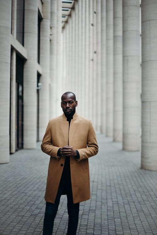 Photo of Man in Brown Coat Posing in the Middle of a Paved Hallway