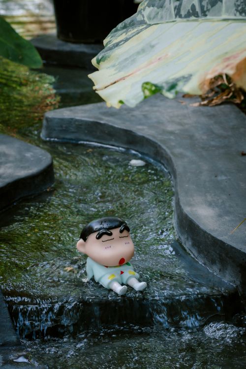 Free A toy character sitting by a flowing garden waterway, surrounded by lush foliage. Stock Photo