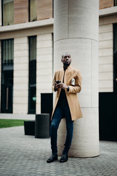 在现代都市中，一个穿着骆驼外套的男人靠在柱子上