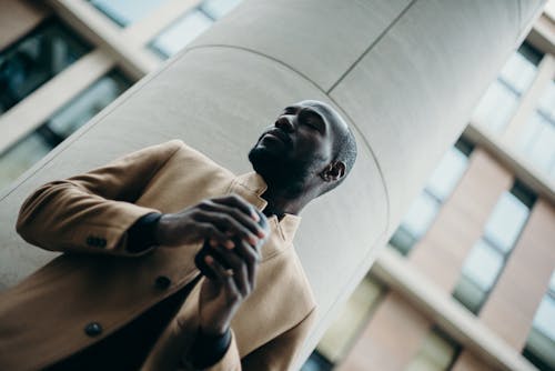 Low Angle Photo of Man in Brown Coat Standing Beside White Column Posing With His Eyes Closed