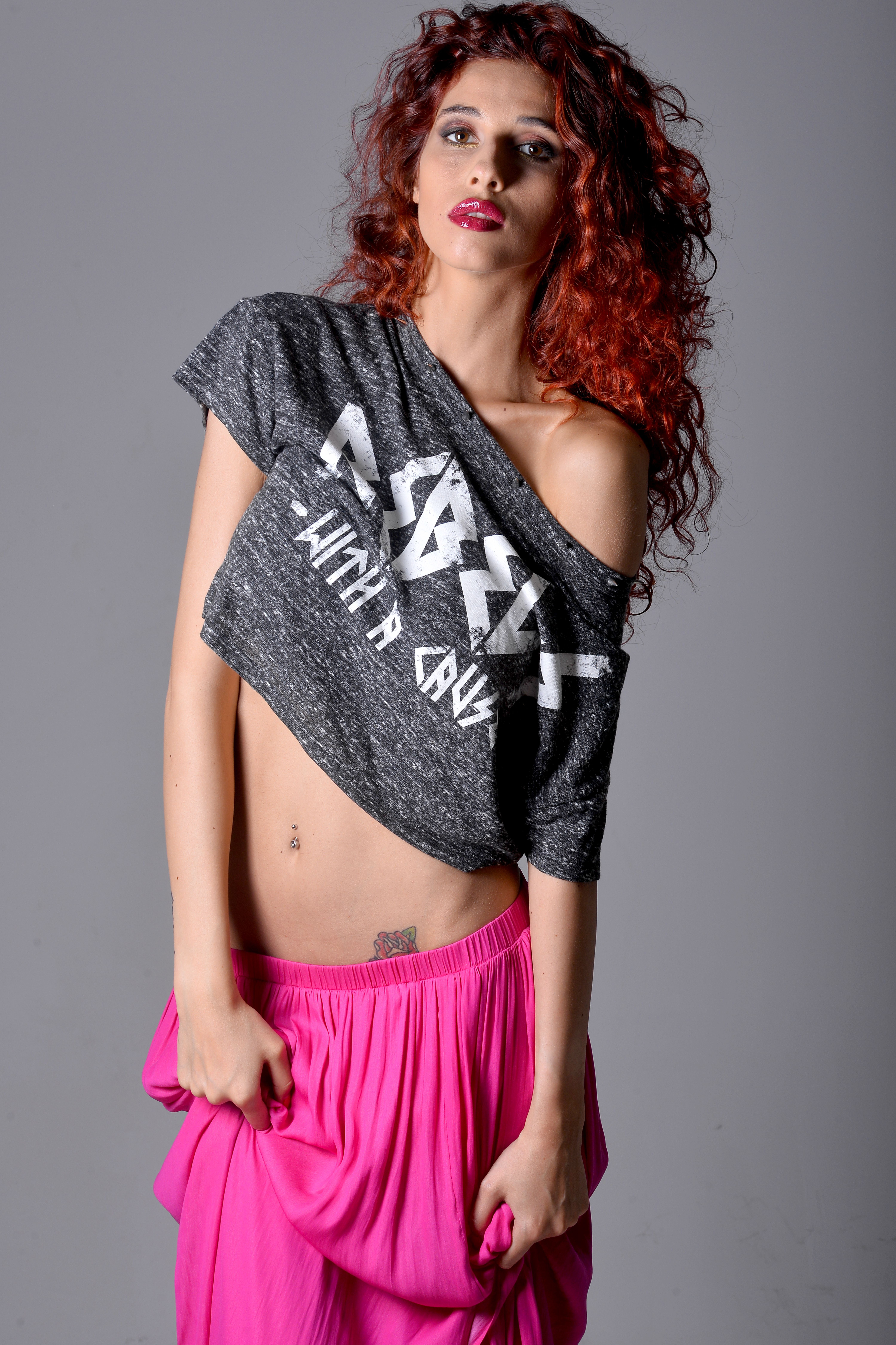Stylish woman with red curls wearing an off-shoulder top and pink skirt, striking a pose.