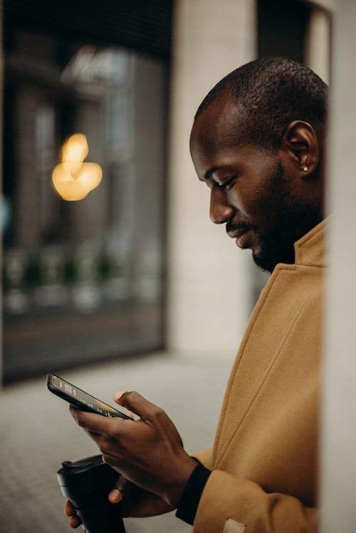 Free Side View Photo of Man in Brown Coat Using Smartphone While Holding Cup of Coffee Stock Photo