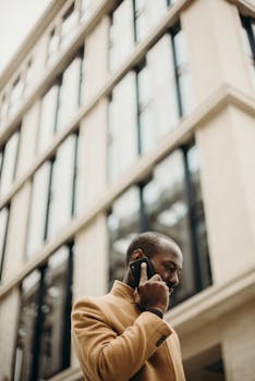 在现代办公楼背景下，一位穿着米色外套的专业男子正用手机聊天