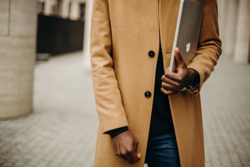 Free Man in a chic tan coat carrying a laptop. Modern fashion on an outdoor street. Stock Photo