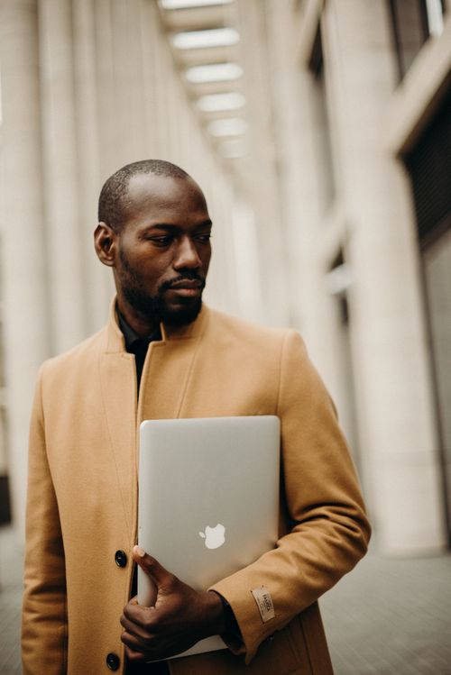 Free A man in a coat holds a laptop, standing thoughtfully in a cityscape. Stock Photo