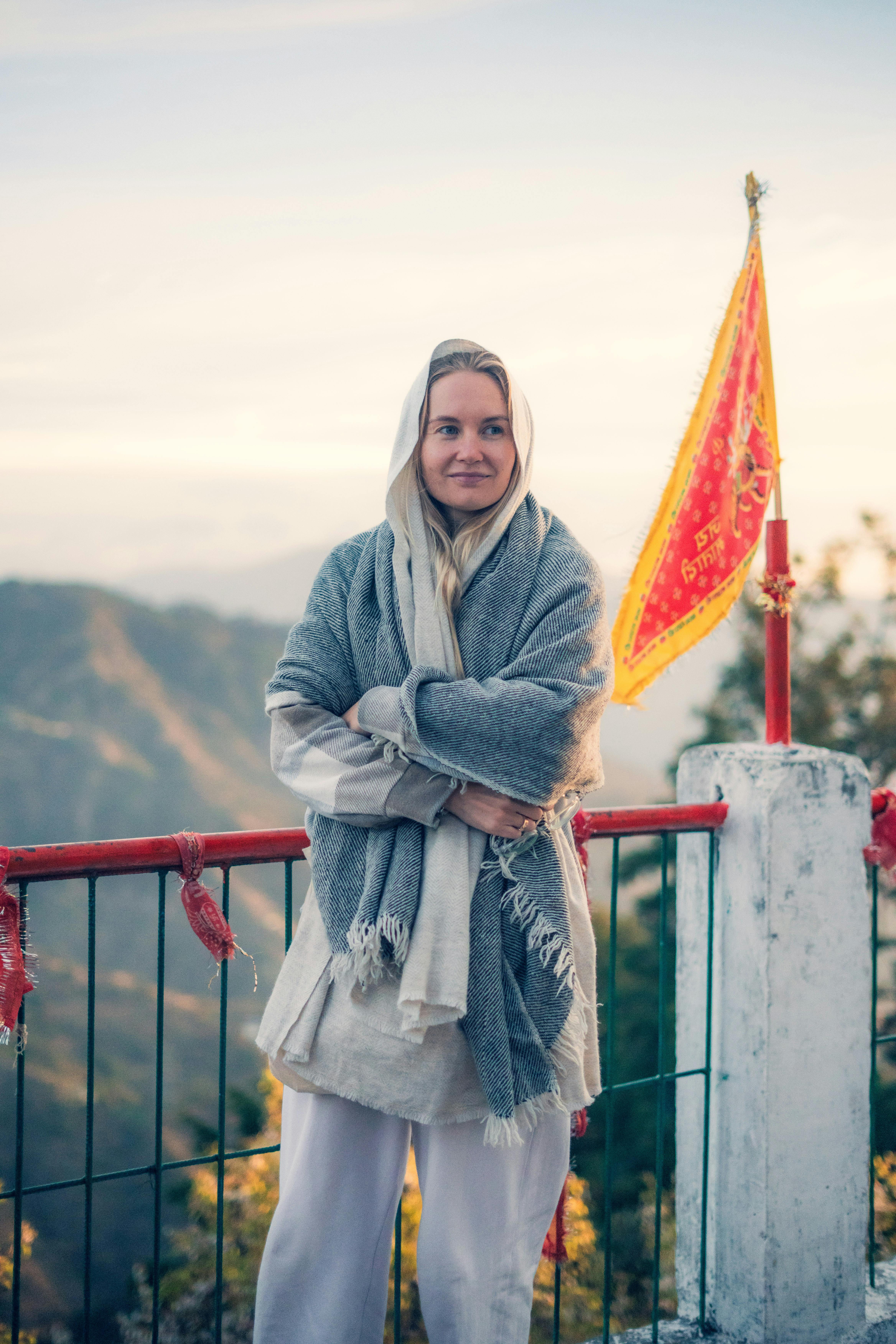 A woman wrapped in a shawl stands on a balcony with mountains in the background.