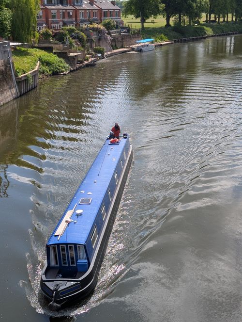 Free A blue narrowboat sails down a peaceful river beside picturesque houses on a sunny day. Stock Photo