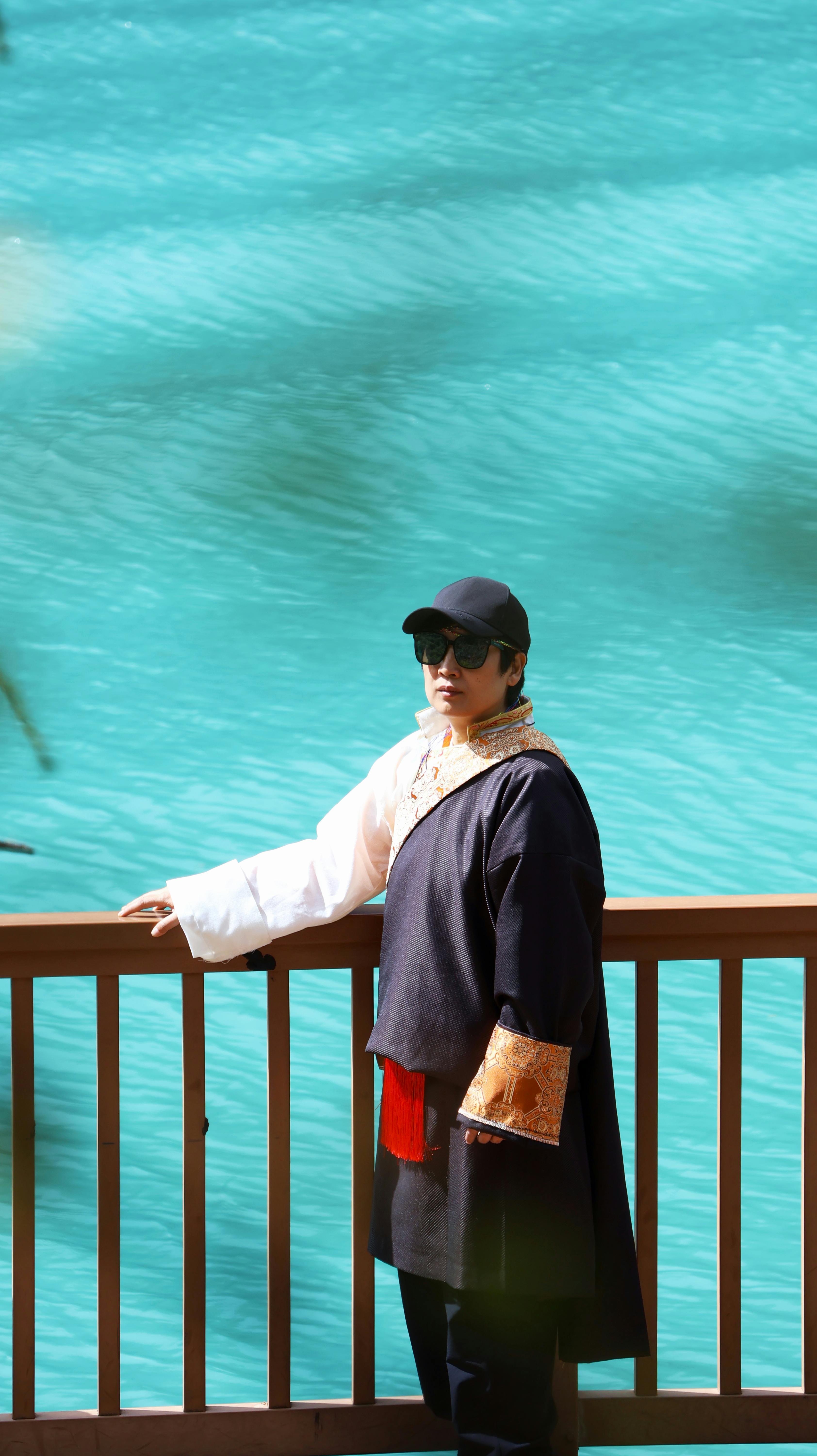 Person in cultural clothing standing by a vibrant lake in Nyingchi, China, exuding serene tranquility.
