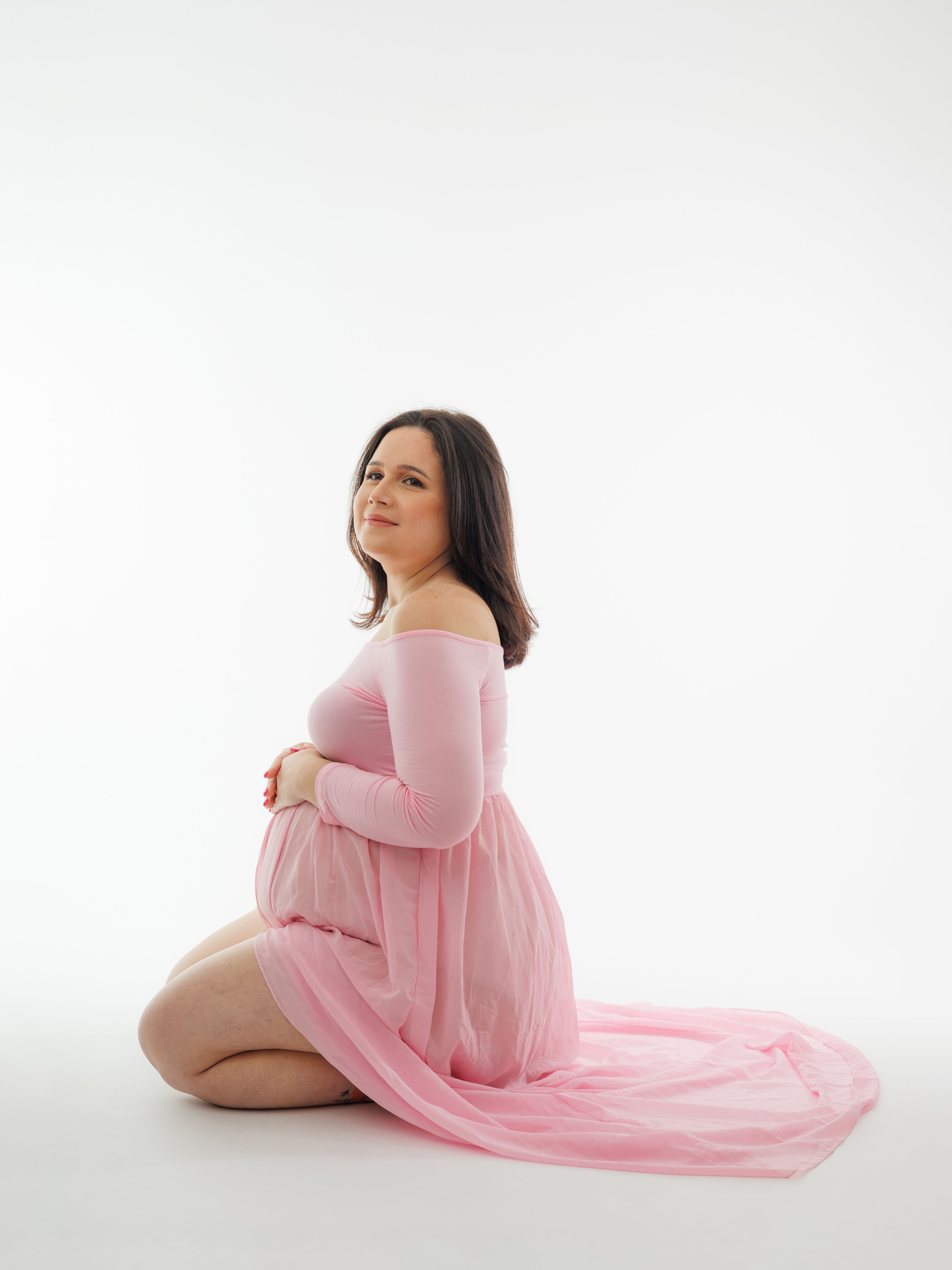 Elegant portrait of a pregnant woman in a flowing pink dress, seated on a white background.