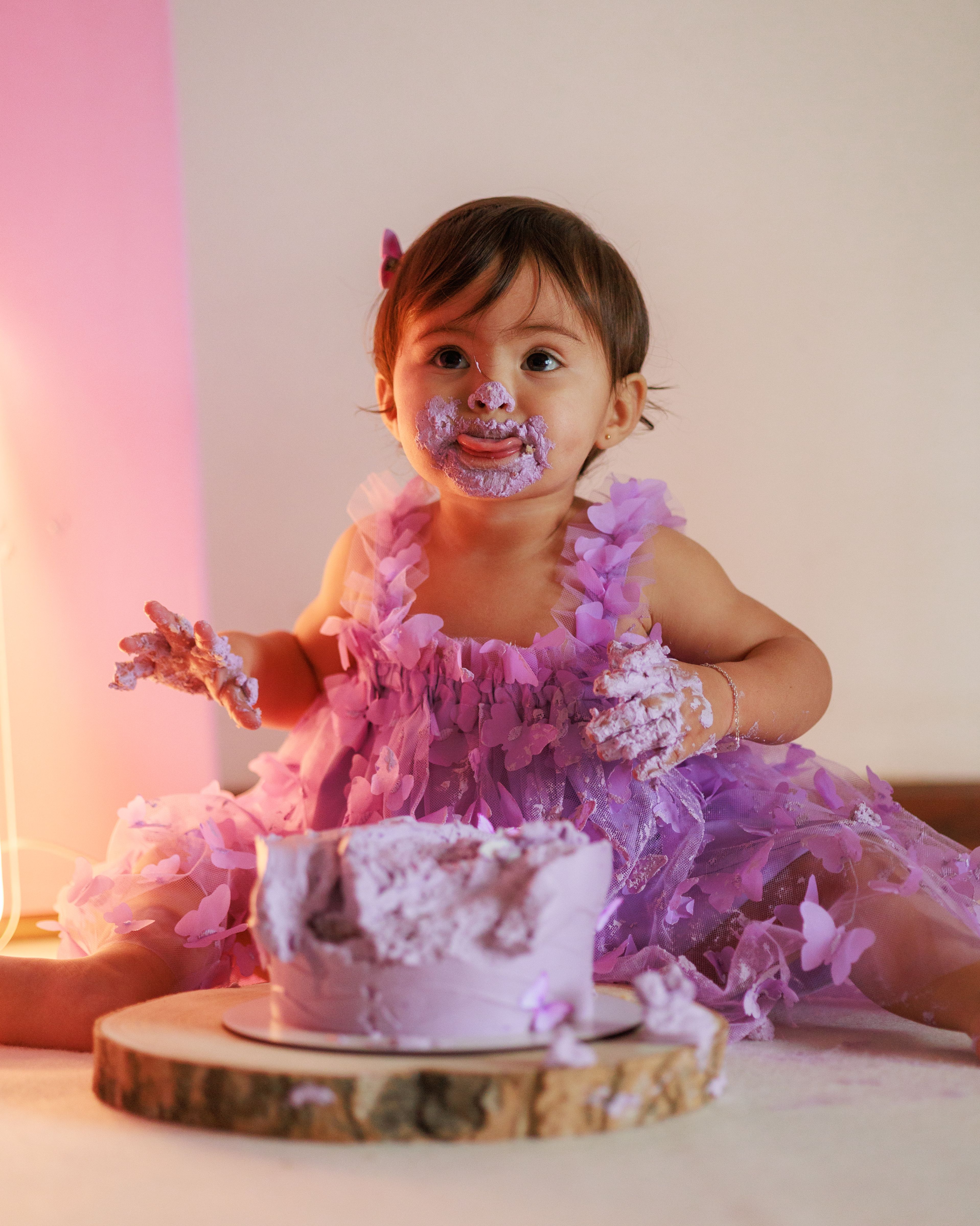 Cute toddler enjoying a messy first birthday cake smash in a purple dress.