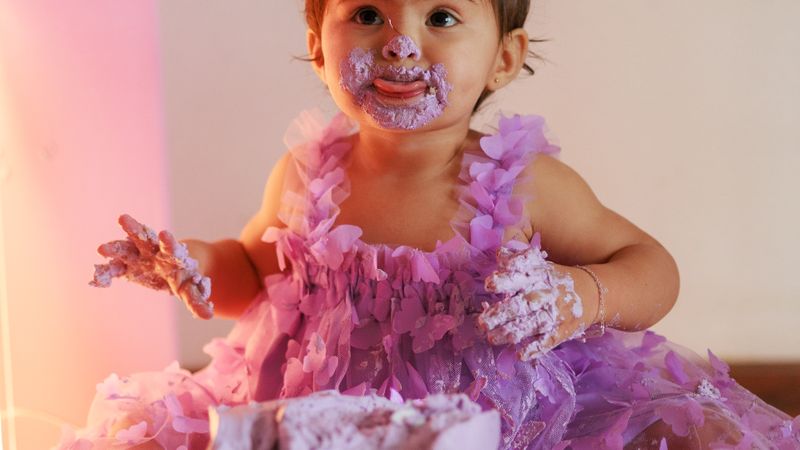 Cute toddler enjoying a messy first birthday cake smash in a purple dress.