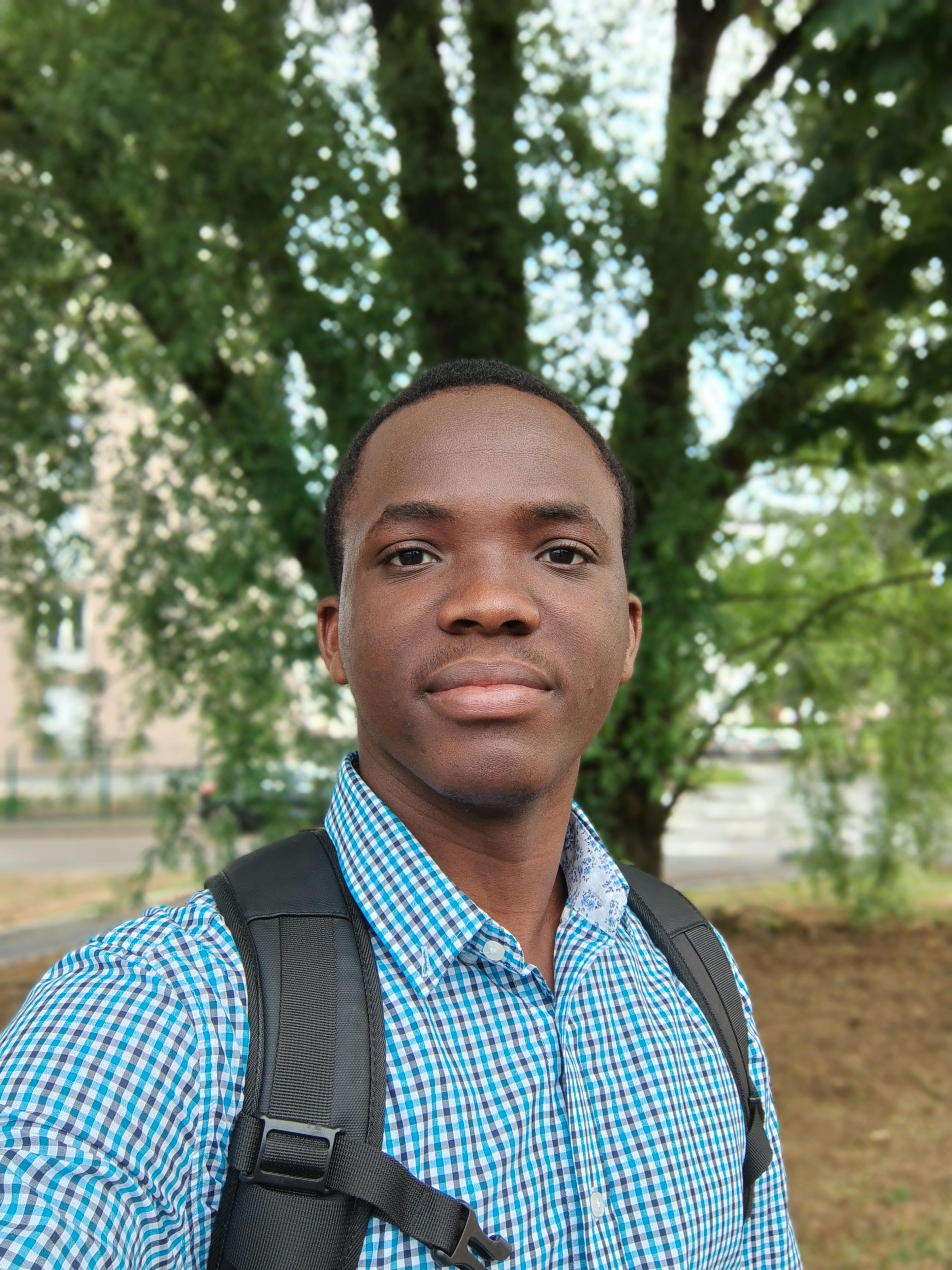 Young adult male taking a selfie outdoors with trees in the background, wearing a checkered shirt and backpack.