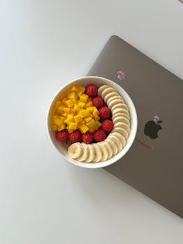 Overhead view of a nutritious fruit bowl with mango, banana, and raspberry slices beside a laptop