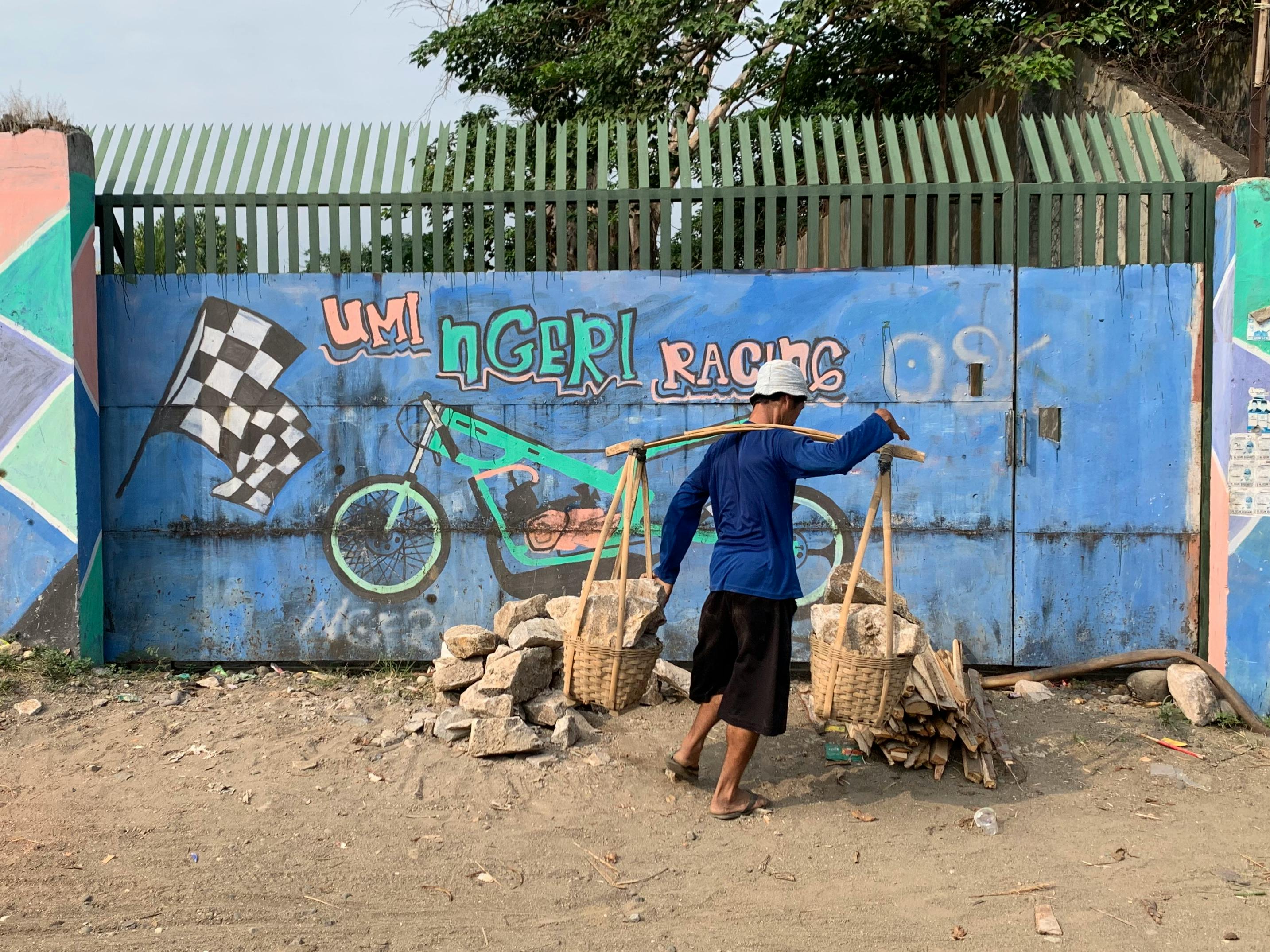 Man carrying baskets in front of vibrant street mural in Sukabumi, Indonesia, depicting daily life.