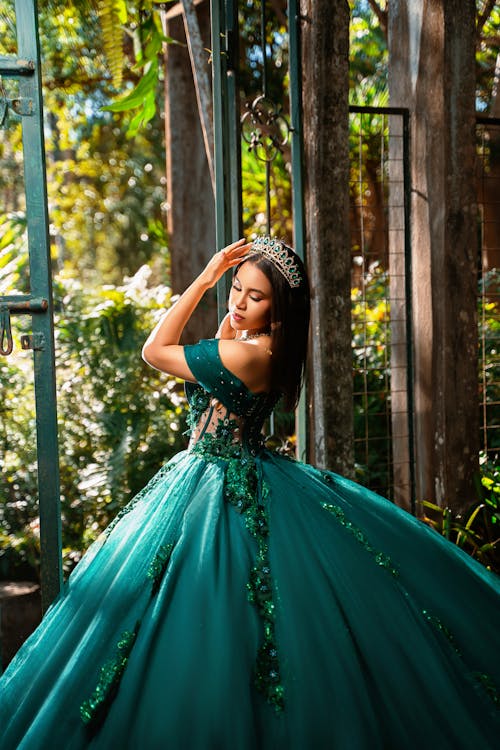 Free A woman elegantly poses outdoors in a stunning green gown and tiara, bathed in sunlight. Stock Photo