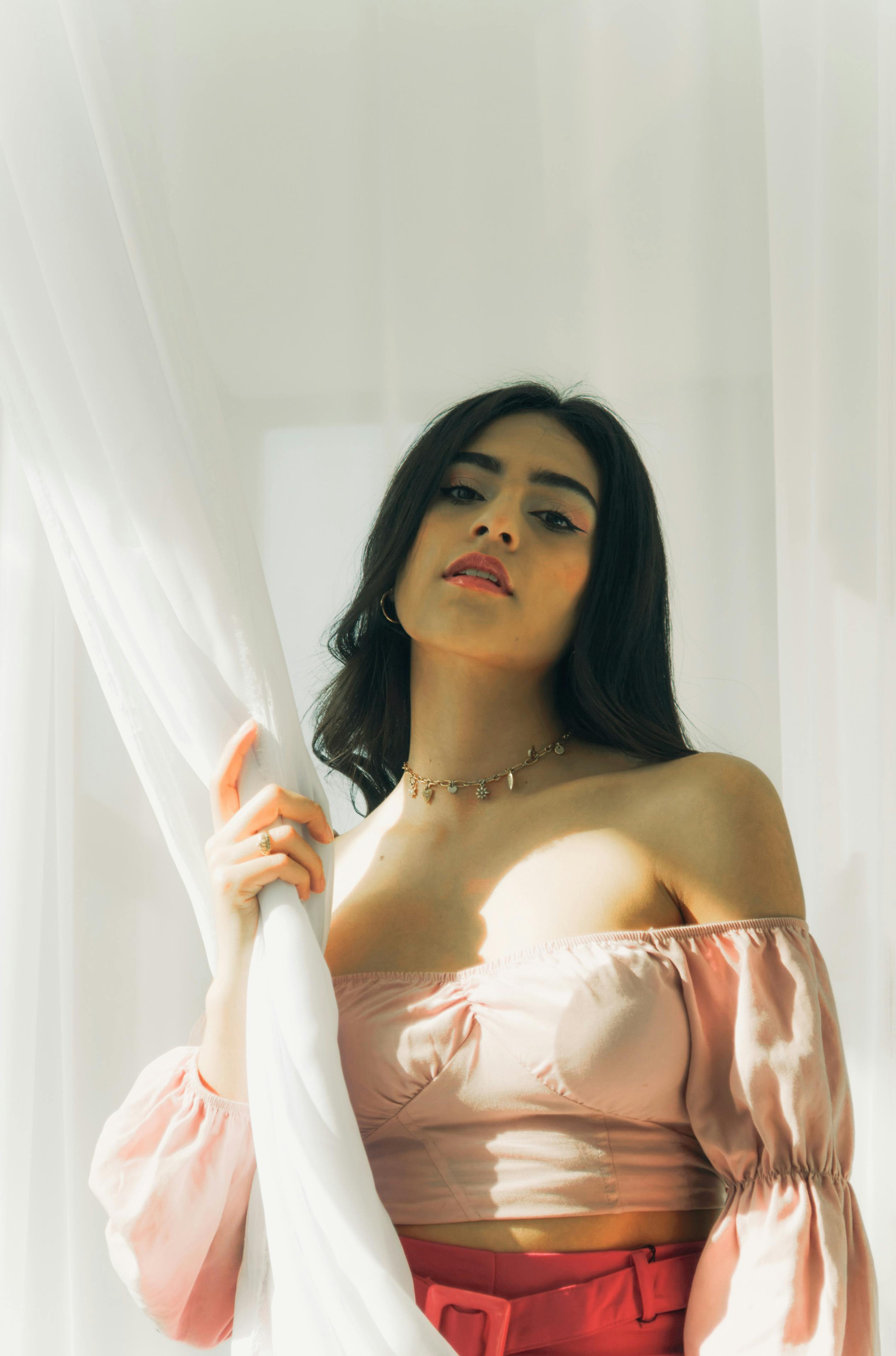 Stylish woman in soft lighting holding a curtain, exuding elegance and confidence.