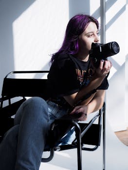 A young woman with purple hair thoughtfully holding a camera in a sunlit studio.