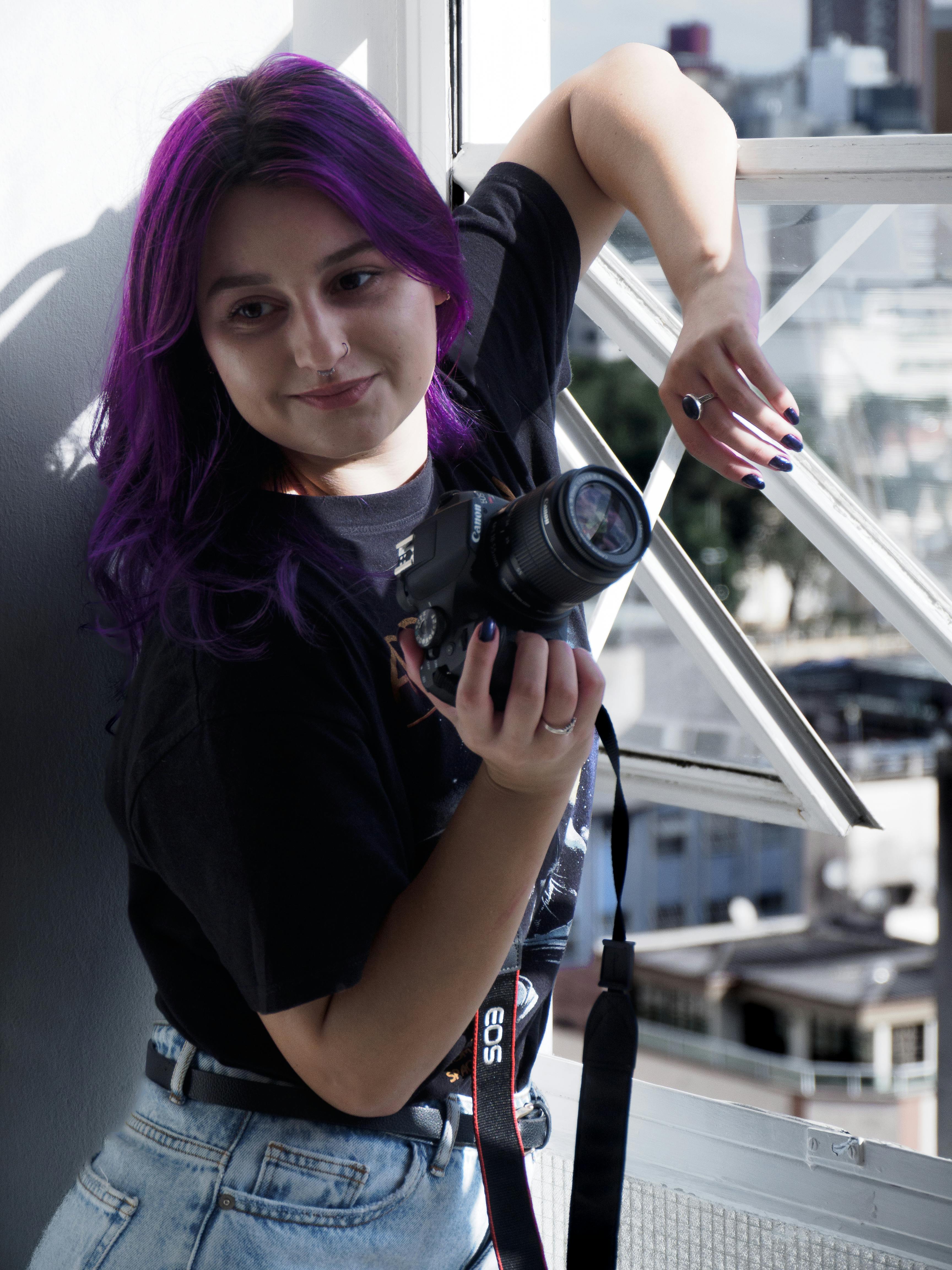 Young woman with purple hair poses with a camera by a sunlit window in the city.