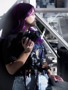 Young woman holding a camera, gazing out a window in an urban environment, with vibrant purple hair.
