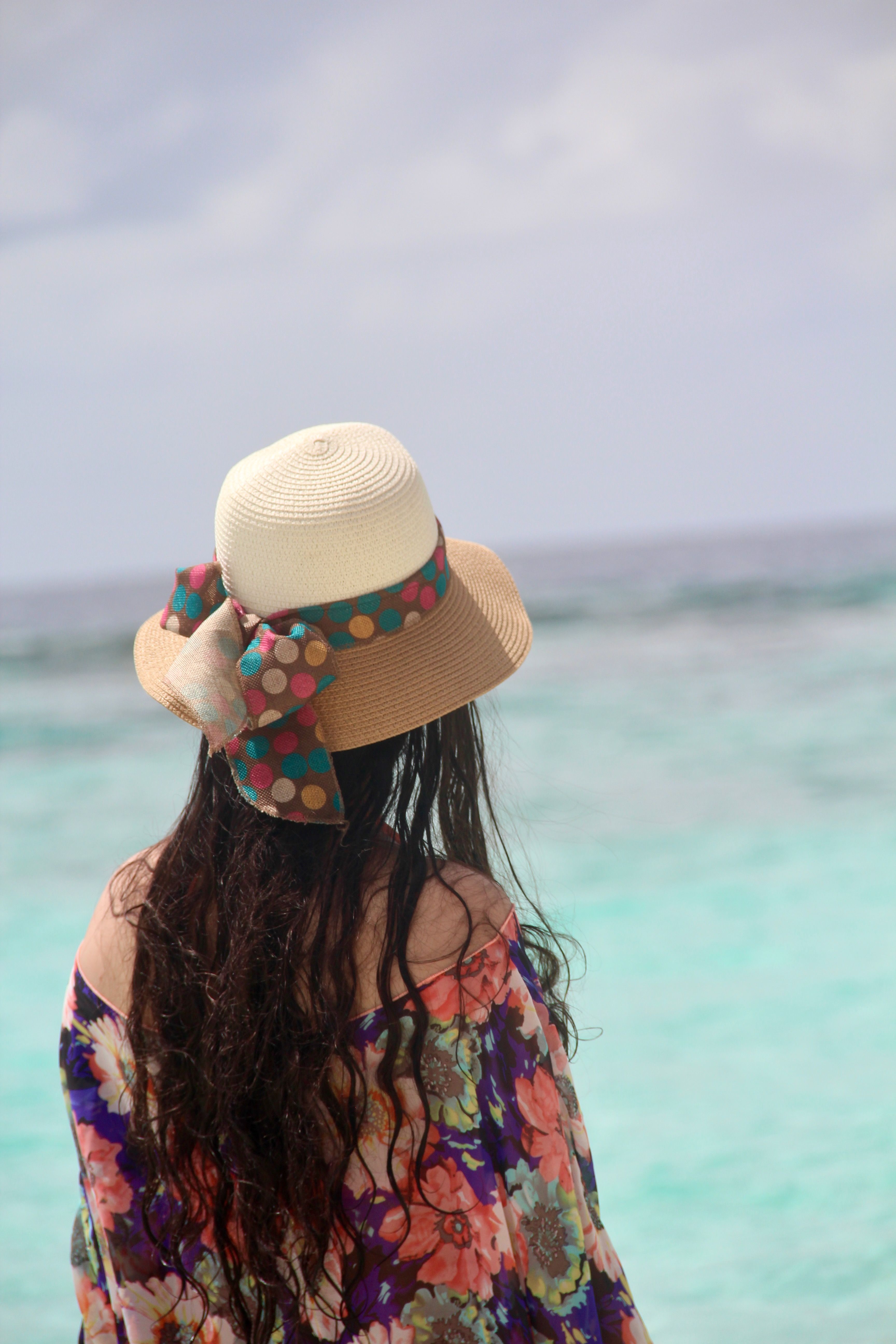 A woman in a colorful floral dress and sun hat looks out at the ocean.