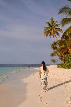 A woman in a floral dress walks along a tropical beach at sunset, surrounded by palm trees.