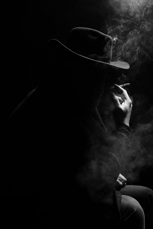 Free A silhouette of a person in a hat smoking, captured in dramatic black and white lighting. Stock Photo