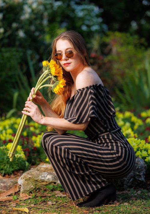 Free Stylish woman in a garden holding sunflowers, wearing sunglasses and a striped dress. Vibrant setting. Stock Photo