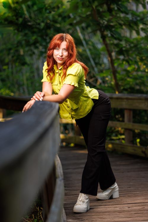 Free Red-haired woman in vibrant outfit posing on a wooden boardwalk surrounded by lush greenery. Stock Photo