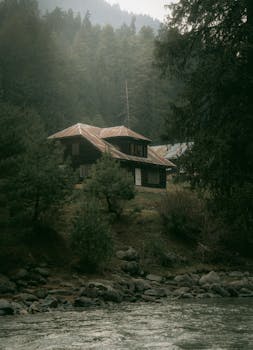 A serene cabin nestled in a misty forest by a river, creating a tranquil and picturesque scene.