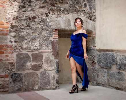 Woman in a stylish blue dress stands confidently against a rustic stone wall.