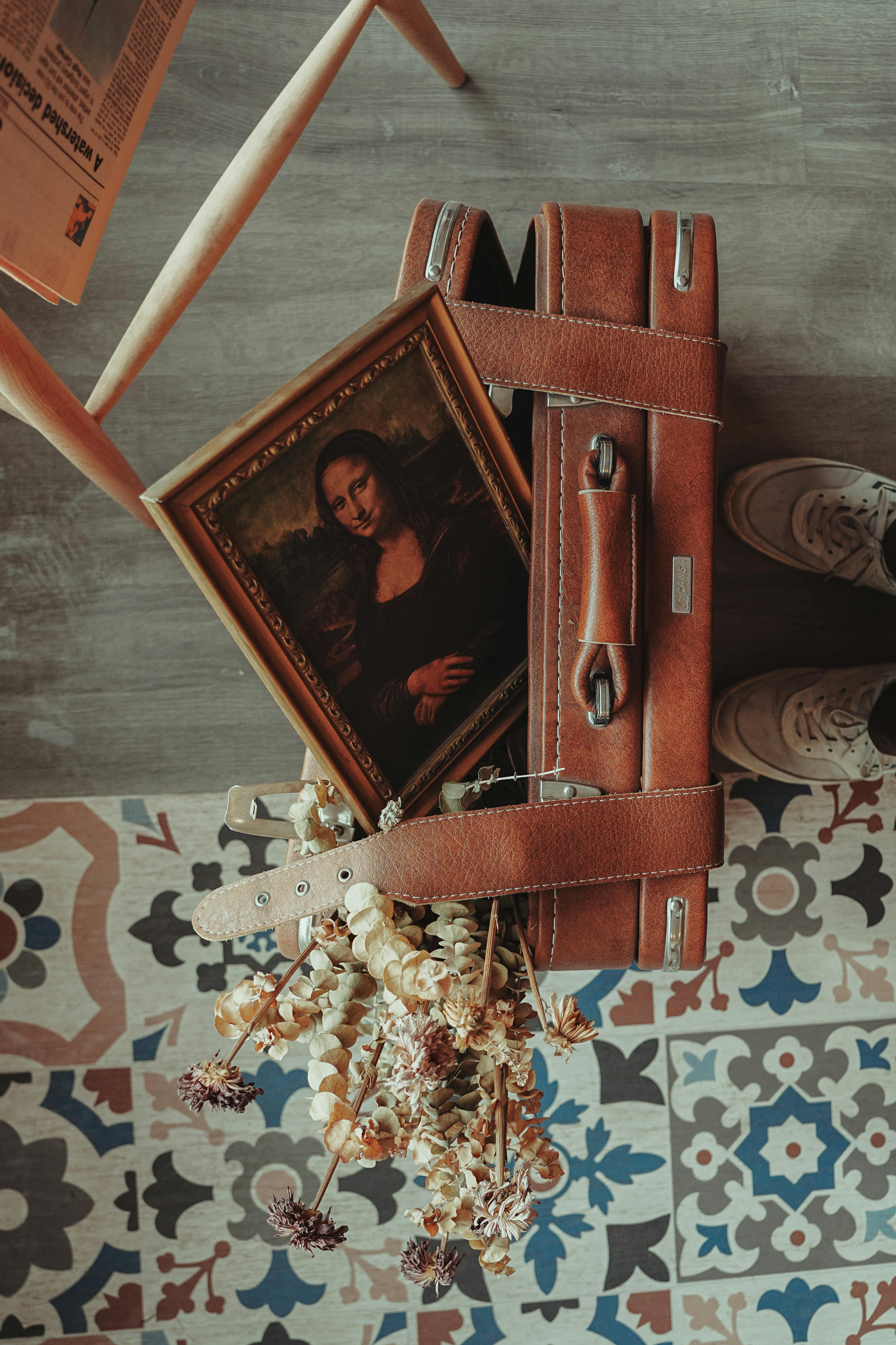A framed Mona Lisa painting atop a vintage suitcase with a floral backdrop on a patterned tiled floor.