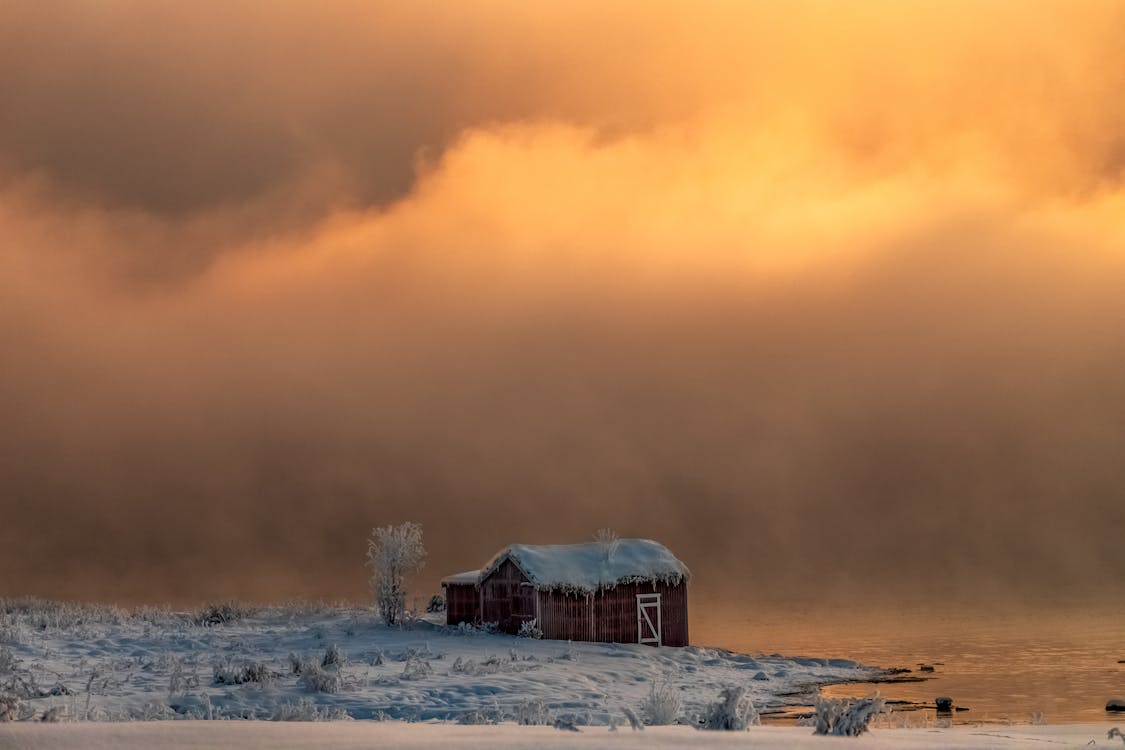 A Brown Barn Close To The River Under Cloudy Skies And Snow Covered Grounds In Winter