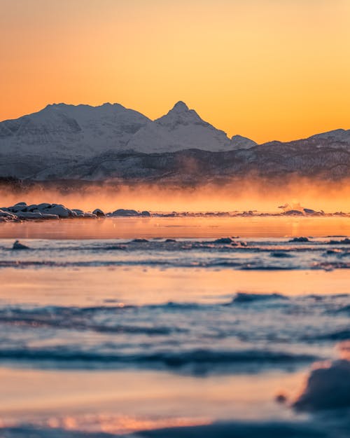 Low Angle Shot Of Snow Covered Ground By A Misty River With The View Of Snow Capped Mountains