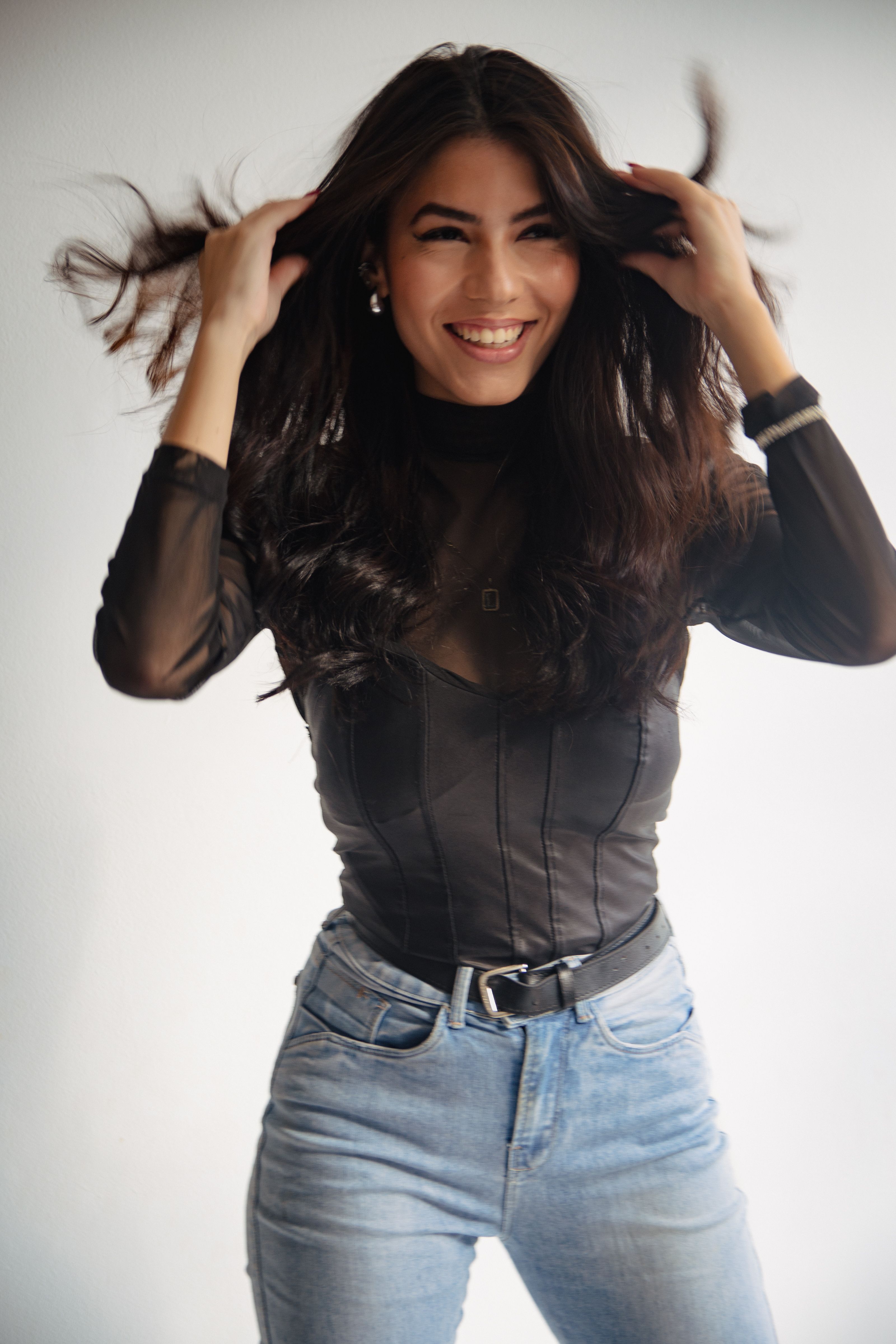 Young woman playfully tossing hair, wearing black sheer top and jeans, smiling happily.