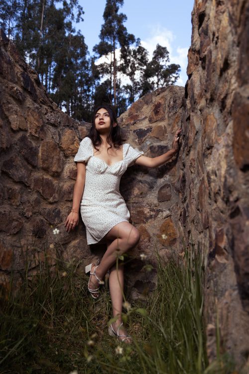 Free Portrait of a woman in a white dress posing in a rustic stone wall setting outdoors. Stock Photo