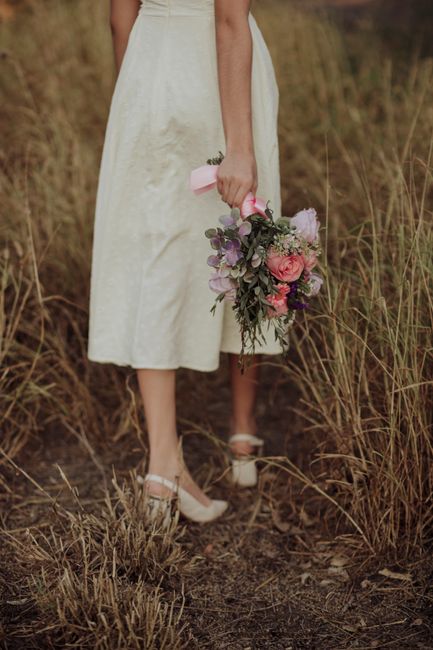 Elegant woman in white dress holding bouquet with pink and purple flowers in grassy field.