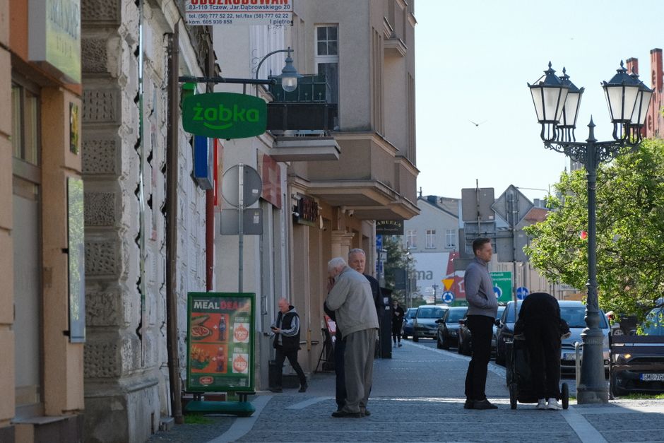 Klient stojący przy kasie w sklepie Żabka, patrzący na kolorowy wyświetlacz jednorazowych e-papierosów, z wyrazem zastanowienia na twarzy.