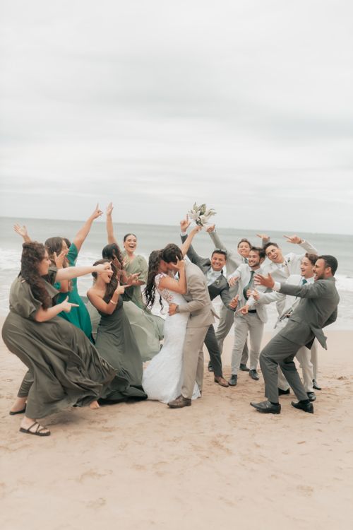 Free Couple kissing on beach surrounded by cheering friends in wedding attire, celebrating their marriage. Stock Photo