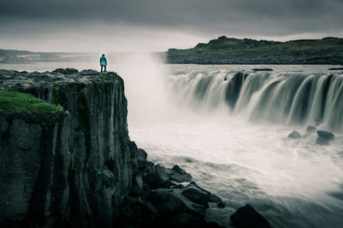 滝の近くの岩の上に立っている青いジャケットの男