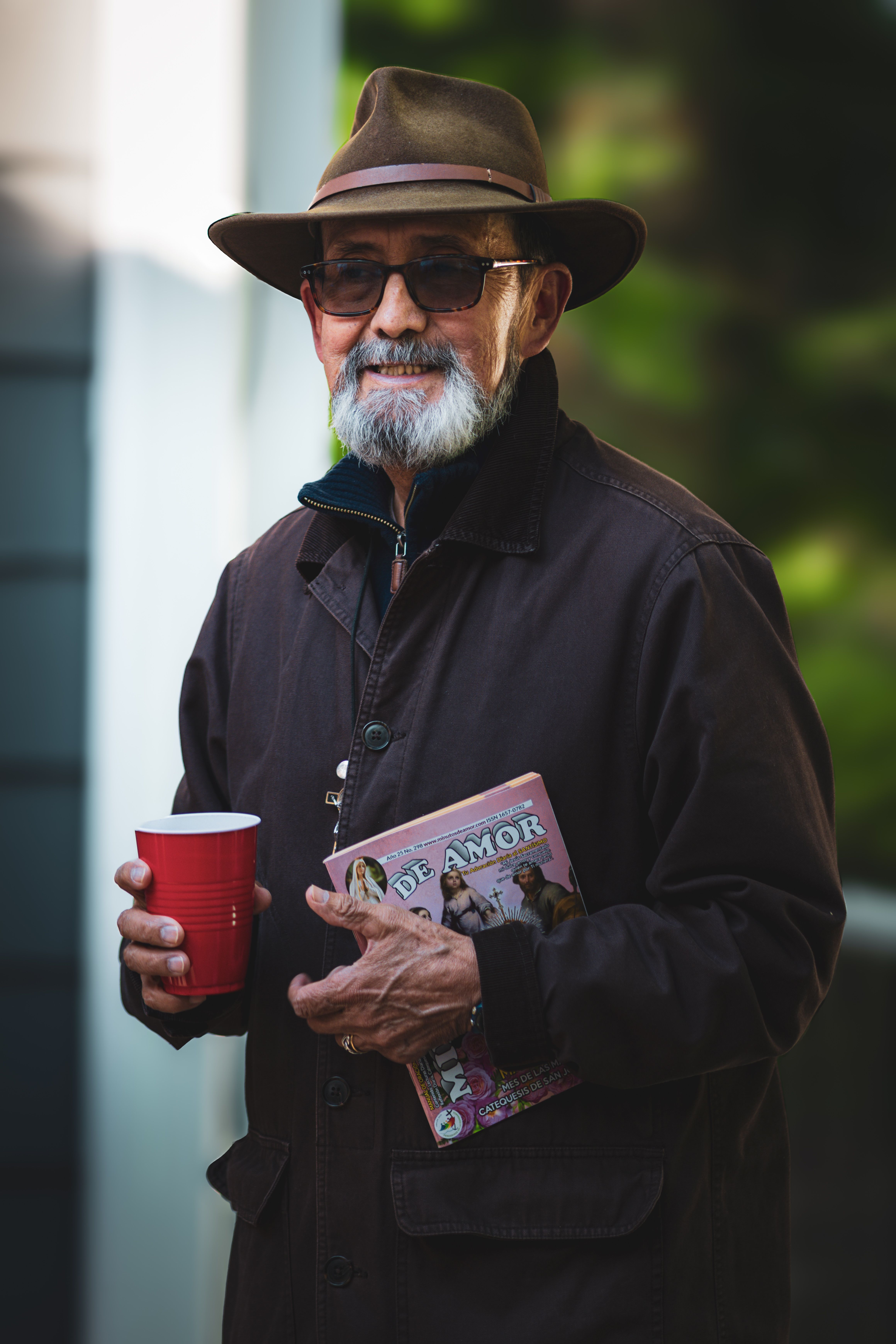 Elderly man in hat and sunglasses holding a magazine and cup, enjoying a relaxed outdoor moment.