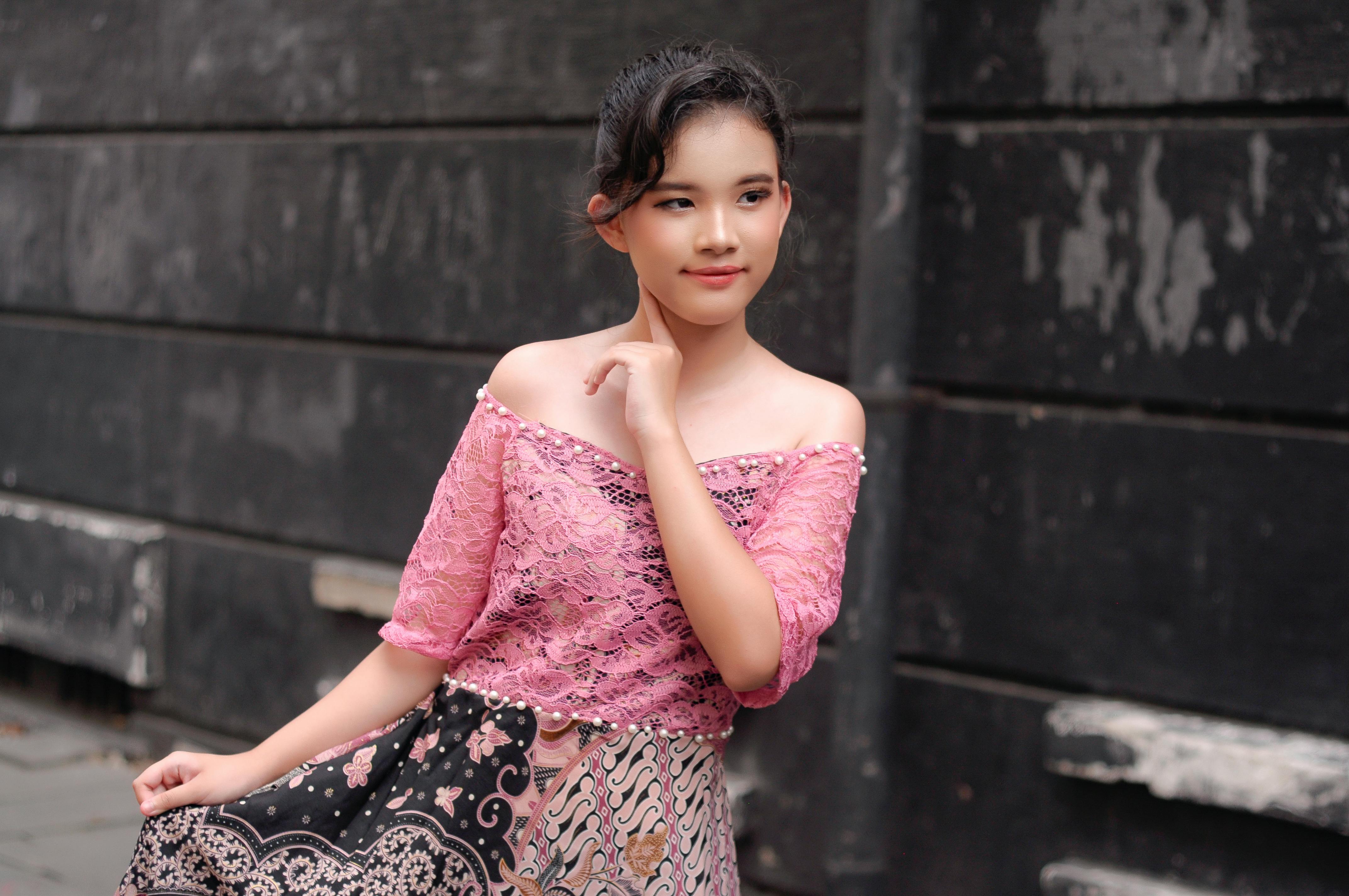 A young woman poses gracefully in traditional attire against a rustic backdrop in Jakarta.
