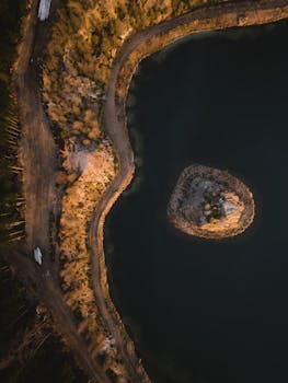 Drone shot of a small island in a tranquil lake surrounded by rugged terrain at sunset.