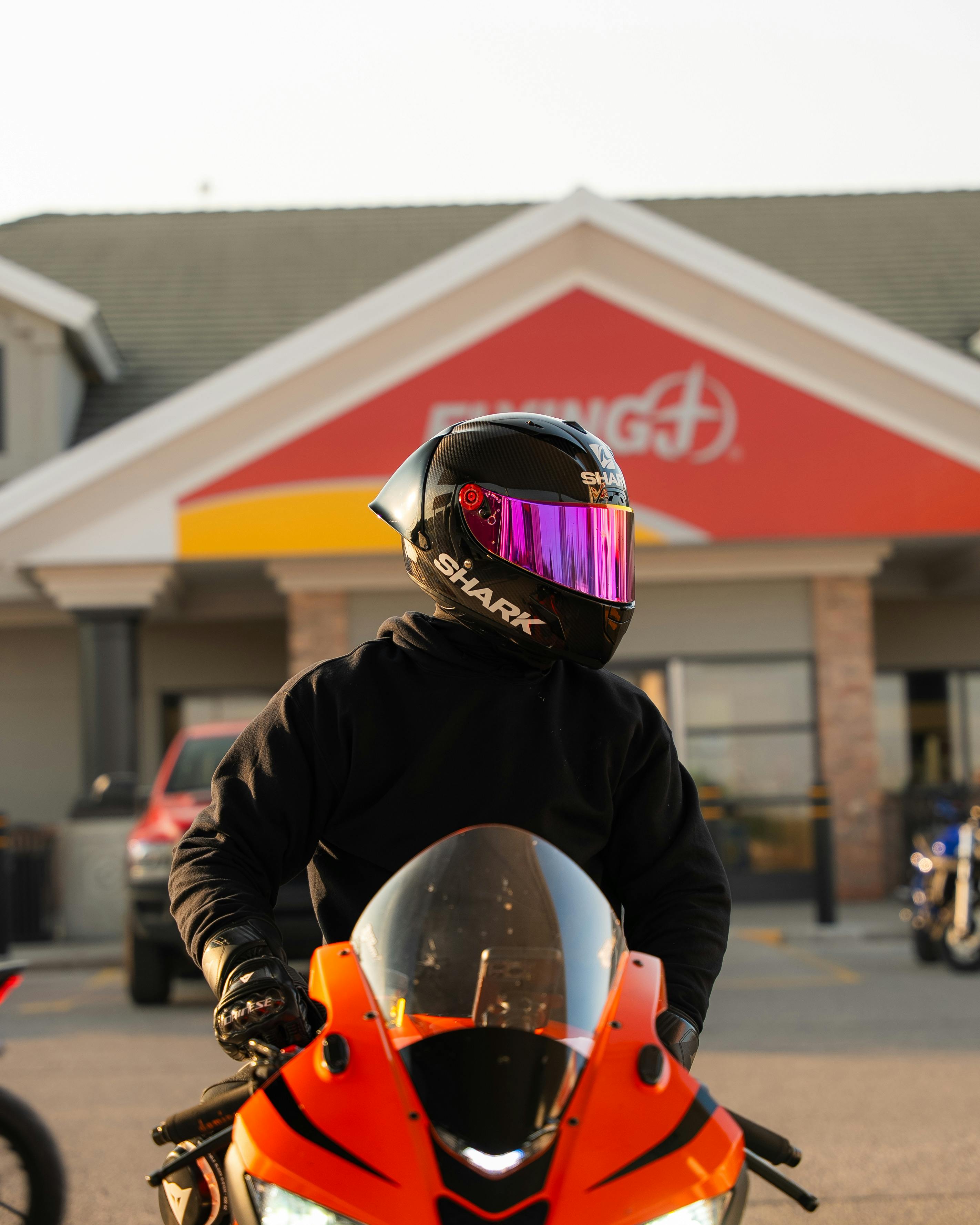 Free A motorcyclist wearing a black helmet with a pink visor stands in front of a commercial building. Stock Photo